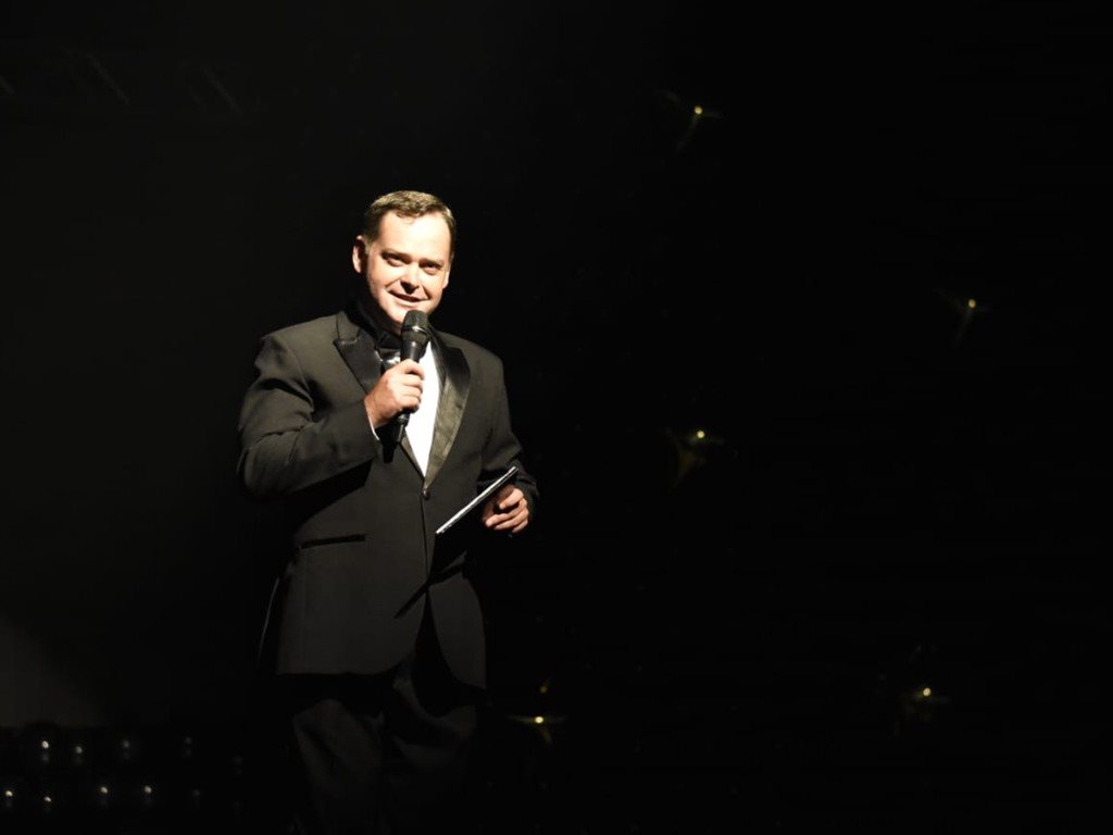 Camilo Lelles apresentando em smoking no palco de evento corporativo