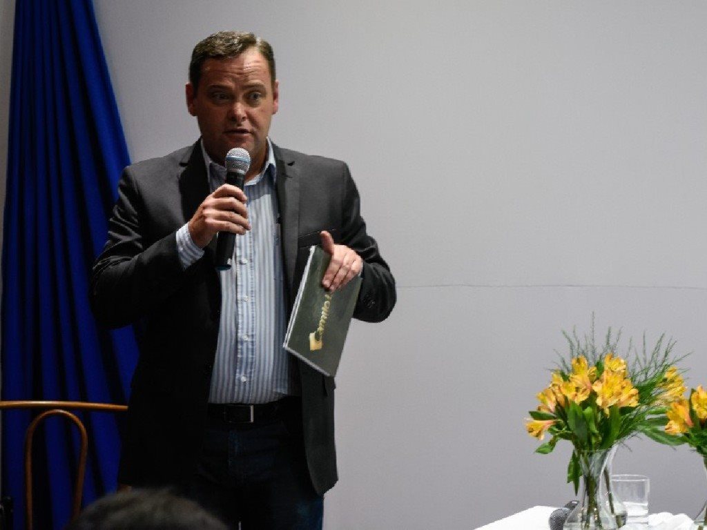 Camilo Lelles como mestre de cerimônia em evento empresarial com pasta e microfone