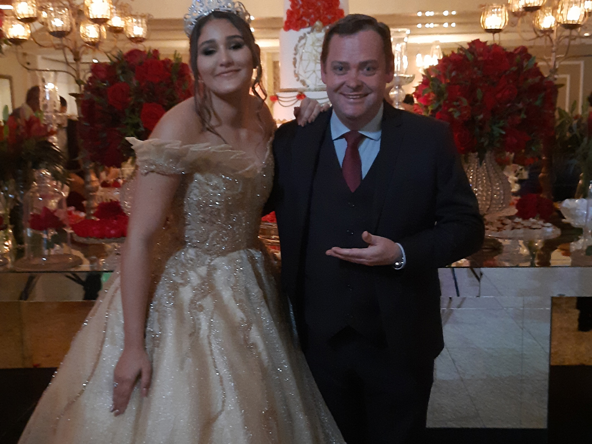 Camilo Lelles com debutante em festa de 15 anos, bolo e flores ao fundo