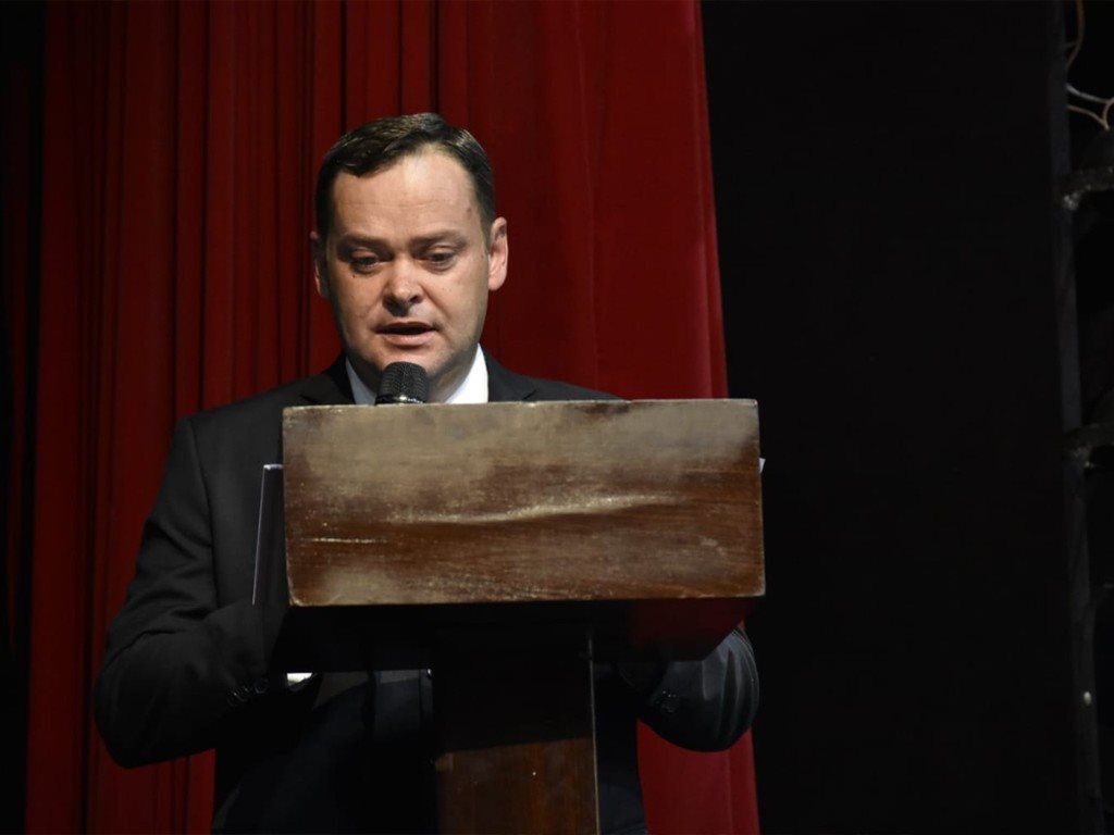 Camilo Lelles como mestre de cerimônia no púlpito de formatura, cortina vermelha ao fundo