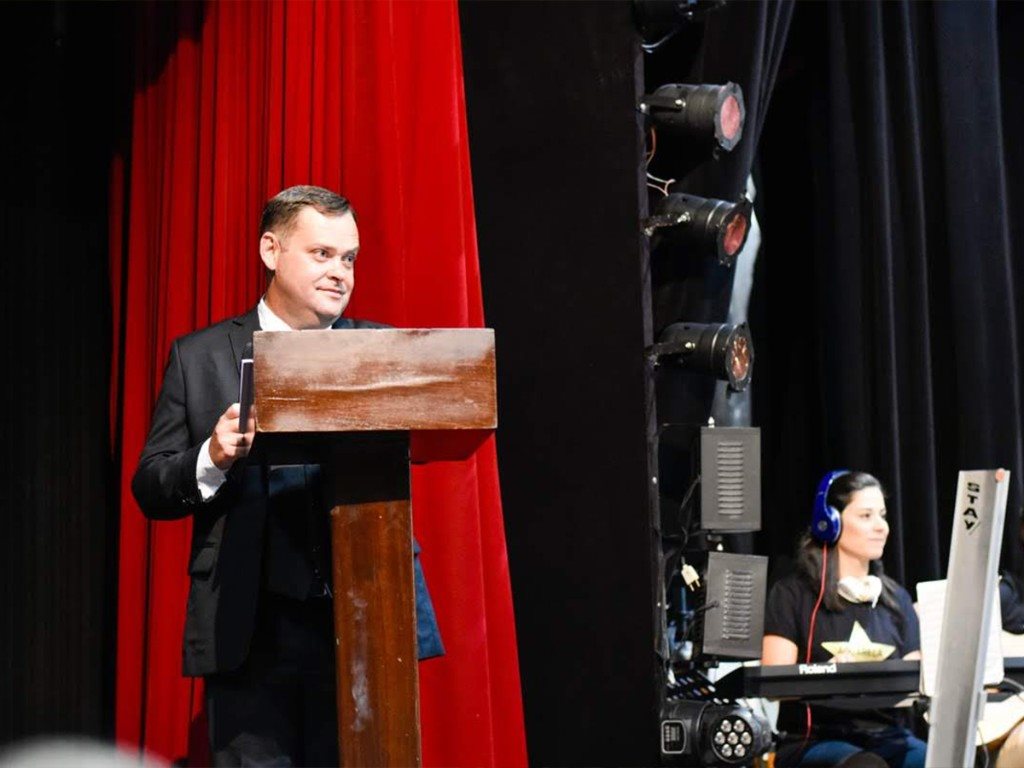 Camilo Lelles no palco de formatura com púlpito de madeira e equipe de som ao fundo